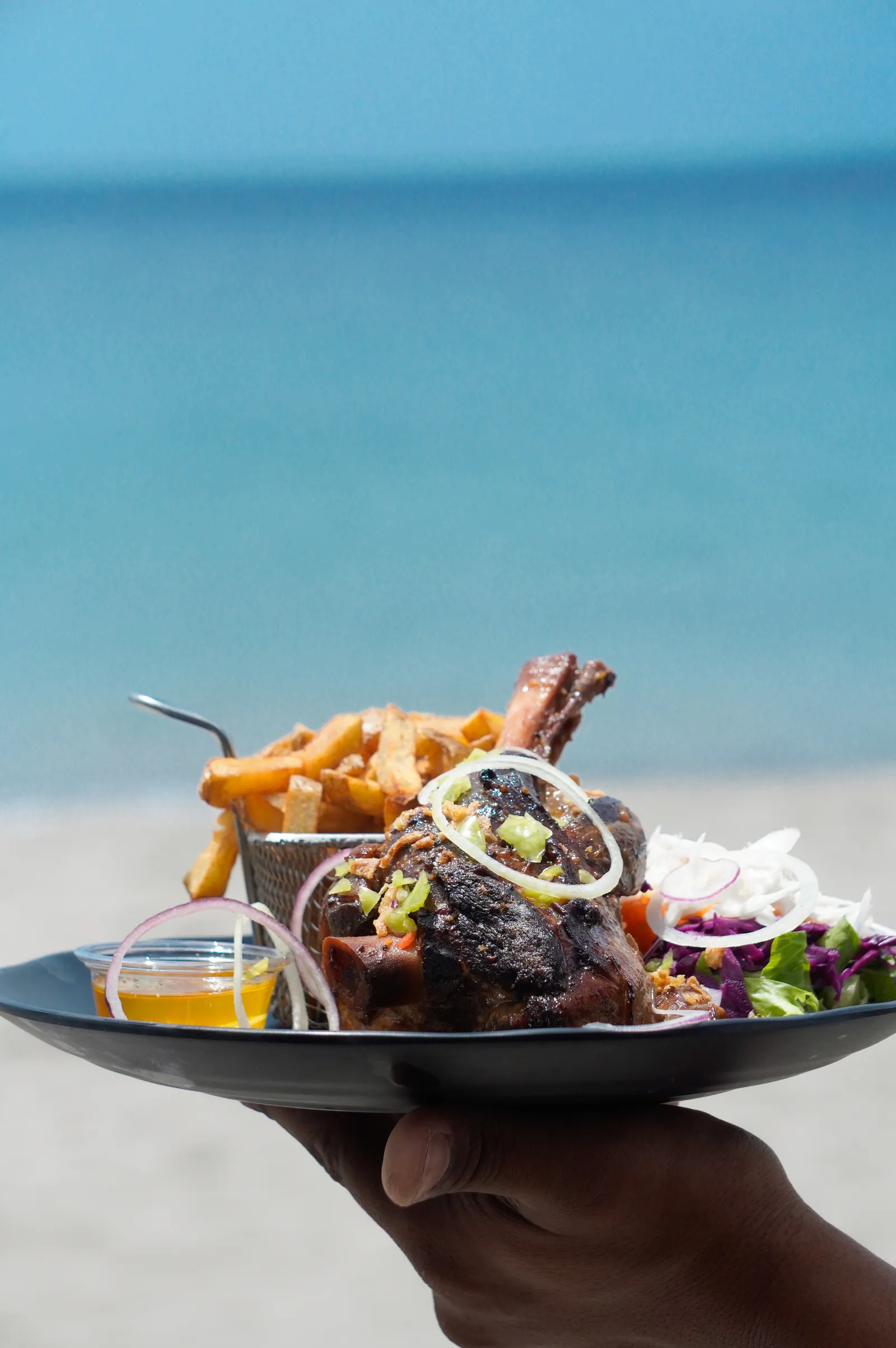 Un serveur présente une souris d'agneau et ses frites maison devant la mer des Caraïbes